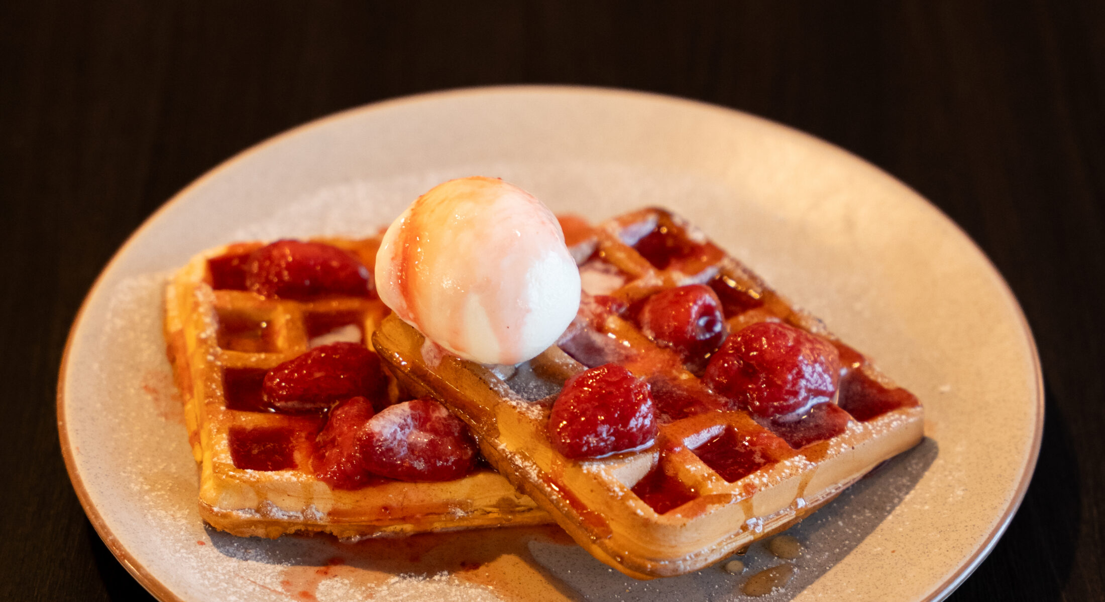 Two golden waffles topped with strawberries, powdered sugar, syrup, and a scoop of vanilla ice cream, served on a light-colored plate.