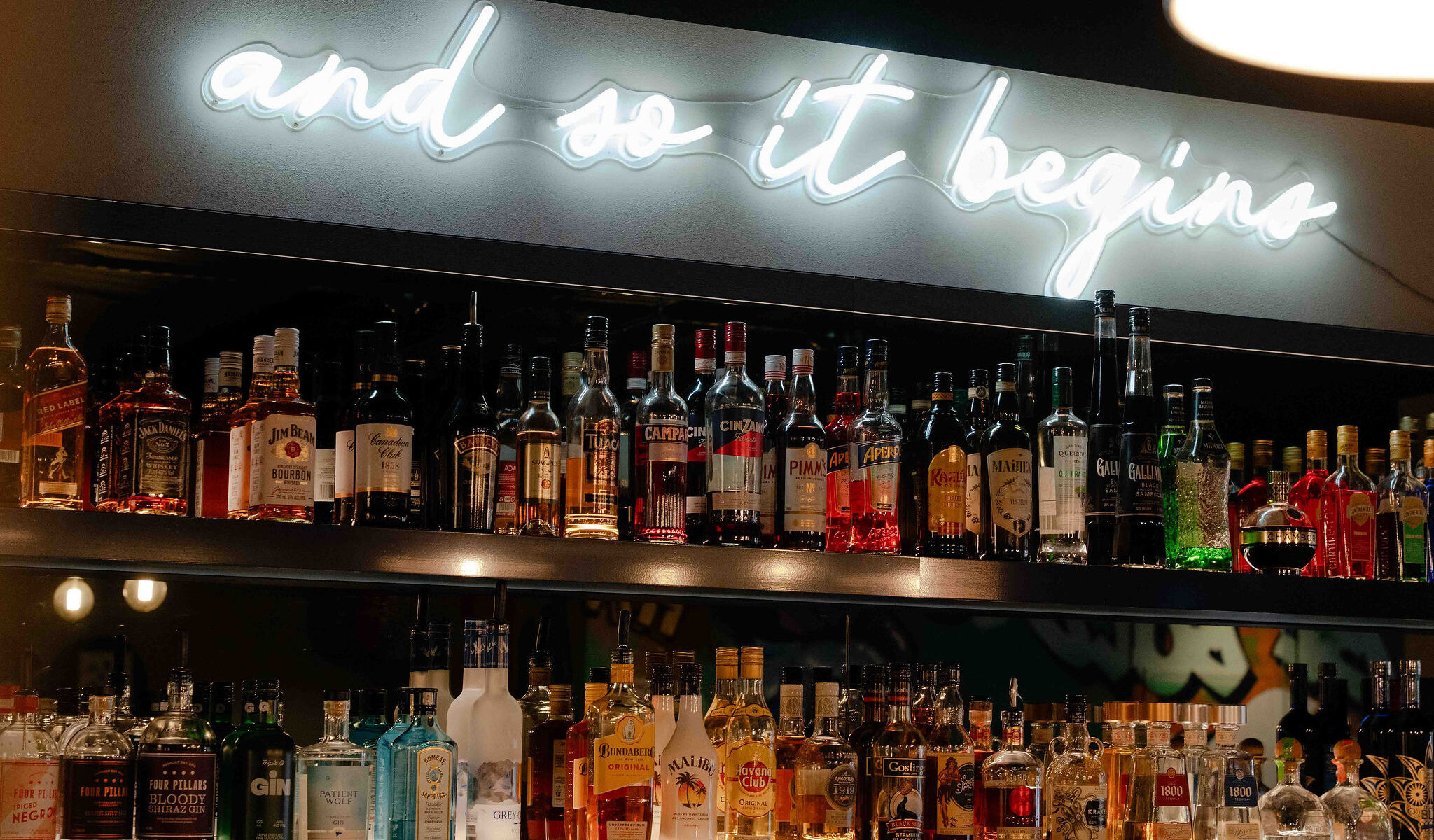 Backlit bar shelves filled with assorted liquor bottles beneath a glowing white neon sign that reads “and so it begins